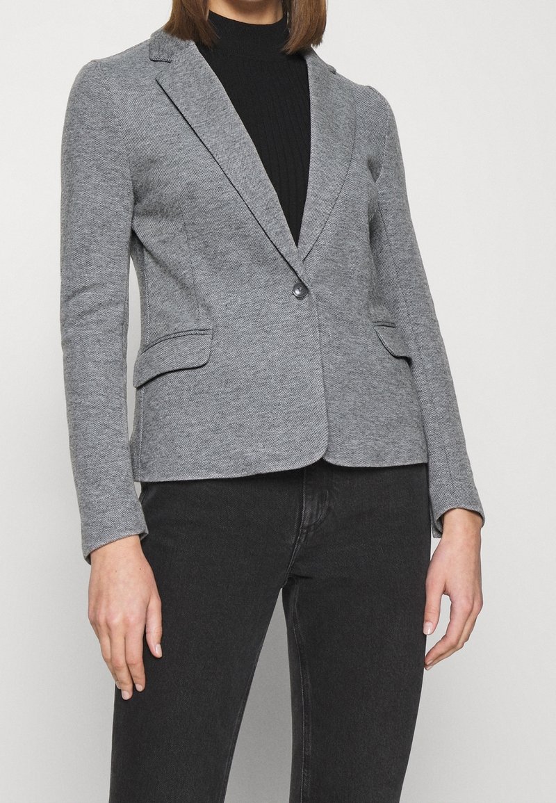 Femme portant un blazer gris cintré sur un col roulé côtelé noir et un jean noir, debout devant un fond blanc uni.