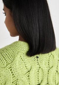 Πράσινη πλεκτή μπλούζα με υφή καλωδίου και μικρό μαύρο κουμπί στο πίσω μέρος, με χοντρή υφή νήματος.