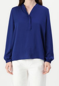 Blaues langärmeliges Oberteil mit V-Ausschnitt und gerafften Manschetten, aus glattem Stoff gefertigt. Kombiniert mit weißen Hosen.