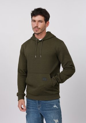 Man draagt een olijfgroen gestructureerde hoodie met voorzak en blauwe gescheurde jeans, staand tegen een effen lichte achtergrond.