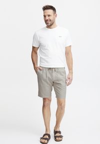 Hvit t-skjorte med korte ermer og tre små fargede prikker på brystet, sammen med beige stripete shorts og brune sandaler.