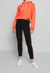 Korálová mikina s bílým šňůrkem, černé džíny s vysokým pasem a knoflíkovou légou a bílé tenisky. Mikina má na boku detaily s visačkou.