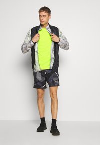 Herrearbedragt sæt: sort og grå mønstret jakke, neon grøn skjorte, sorte geometriske shorts, sorte sportssko og sorte strømper.