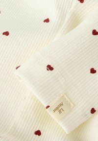 Jasna tkanina z czerwonymi motywami serc; złożenie odsłania metkę z napisem "Lil' Agender", podkreślającą miękkość i zabawny design.