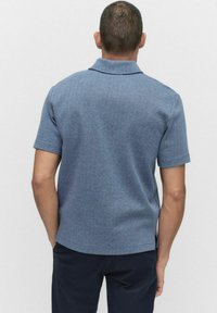 Kortärmad pikétröja i texturerad blått tyg med ett subtilt fiskbensmönster och en vikkrage. Inga synliga logotyper eller detaljer.