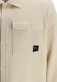 Chemise beige texturée à boutons, col, poche avant à rabat fermée par un bouton, et petite étiquette noire avec un dessin blanc sur le coin de la poche.