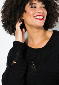 Schwarzer Strickpullover mit geripptem Muster, weitem Ausschnitt und ausgefransten Akzenten an den Ärmeln, mit lockerer Passform. Ausschnittdetails sichtbar.