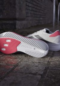 Hvide og lyserøde Adidas Adizero løbesko på brostensbelagt gade, hvor den ene viser den teksturerede sål for greb og støtte.