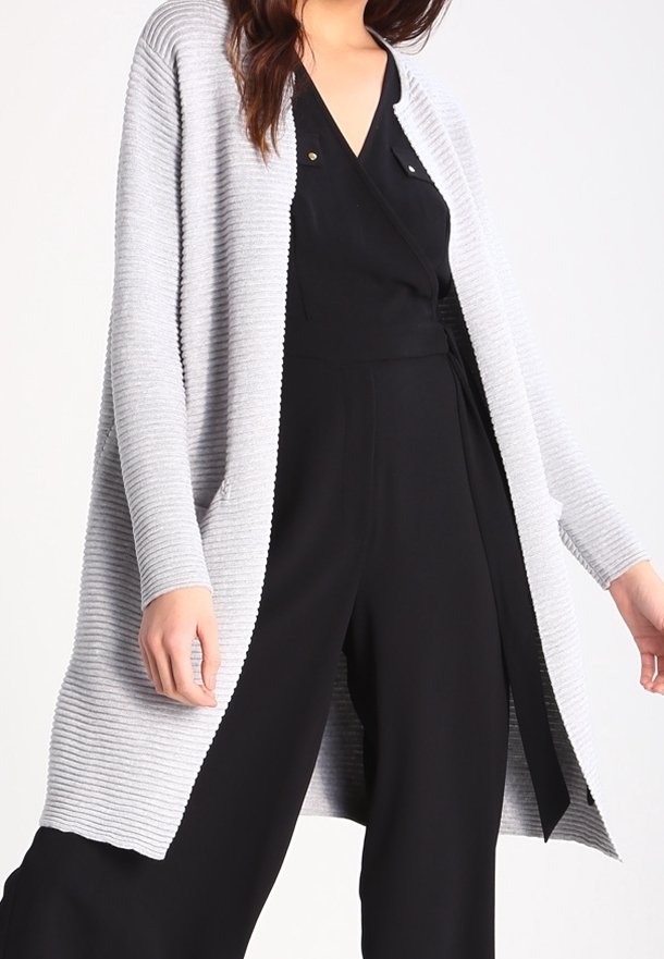 Ljusgrå ribbad cardigan med öppen front, långa ärmar och sidofickor, lagd över en svart jumpsuit med en slät tygtextur.