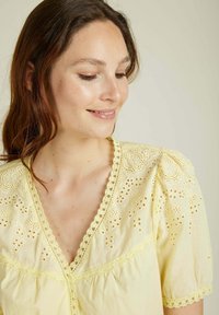 Chemise jaune clair à manches courtes bouffantes, ornée de broderies à oeillets et de dentelle festonnée le long du décolleté et de l'ourlet. Tissu à la texture douce.