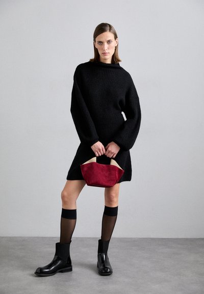 Vestido suéter oversized preto, botas de tornozelo pretas, meias pretas transparentes, segurando uma bolsa de suede vermelha com acabamentos em bege. Fundo simples.
