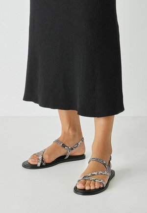 Fødder iført sorte flade sandaler med grå slangeprint krydsede remme, kombineret med en sort ribstrikket midi-nederdel på hvidt gulv.