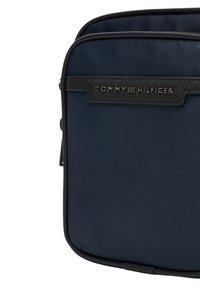 Námořnicky modrá taška s hladkou texturou, černými detaily a výrazným logem TOMMY HILFIGER. Obdélníkový tvar se zavíráním na zip.