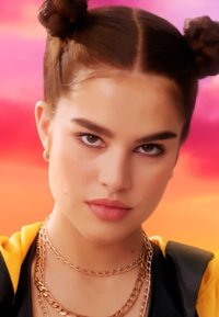 Mujer joven con cabello castaño en dos moños, vestida con cadenas de oro en capas y un atuendo amarillo y negro, contra un fondo vibrante de color rosa y naranja.