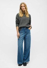 Mujer con cabello largo y rubio, vistiendo un suéter gris, jeans anchos azul, zapatos negros y una pulsera negra, de pie contra un fondo blanco liso.
