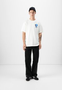Hvid bomulds T-shirt med blåt grafik på venstre bryst, parret med sorte brede jeans og sorte sneakers med hvide striber.