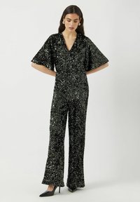 Tuta nera di paillettes con maniche ampie e scollo a V. Presenta una silhouette aderente e pantaloni svasati, abbinata a tacchi alti neri.