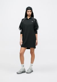 Μαύρο πόλο φόρεμα με λευκές ρίγες στα μανίκια, κατασκευασμένο από βαμβάκι. Συνδυασμένο με γκρι αθλητικά παπούτσια και ανοιχτόχρωμες κάλτσες. Ρυθμιζόμενο καπέλο.