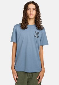 Ung person med langt krøllet hår iført en blå t-shirt med teksten "RIDE THE RAILS" og grønne bukser foran en ensfarvet baggrund.
