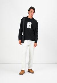 Zwarte lange mouw t-shirt met een graphic op de voorkant en tekst op de mouw, gecombineerd met witte broek en bruine sneakers. Tas over het lichaam gedragen.