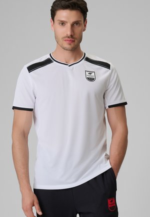 Mężczyzna w białej sportowej koszulce z krótkim rękawem z czarnymi paskami na ramionach i logo oraz czarnych spodniach z czerwonym logo.