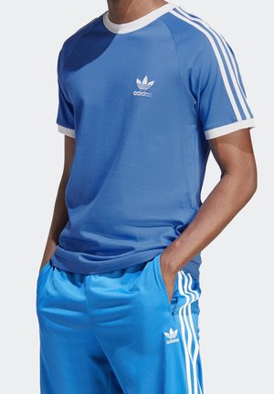 Mann trägt blaues Adidas T-Shirt mit weißen Streifen und Logo, kombiniert mit passenden blauen Adidas Trainingshosen mit weißen Streifen und Logo. Hände in den Taschen.
