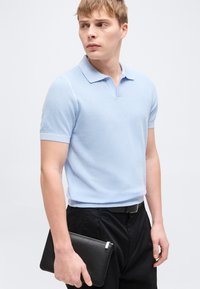 Νεαρός άνδρας με ανοιχτό μπλε κοντομάνικο πουκάμισο πόλο και μαύρο παντελόνι κρατάει μαύρο τσαντάκι, κοιτάζοντας πλάγια σε απλό φόντο.