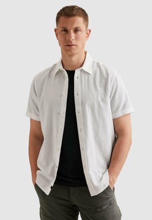 Muž oblečený v rozpnutom bielom tričku s krátkym rukávom cez čierne tričko a zelené cargo nohavice, stojaci s rukami v vreckách.