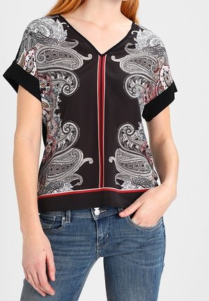 Frau trägt schwarze Kurzarmbluse mit weißen Paisley-Mustern, roten vertikalen Streifen und blauen Jeans, Hand in der Tasche.