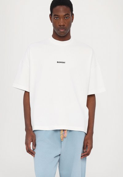 T-shirt blanc surdimensionné avec un col rond, mettant en avant un graphique noir "Bonsai" sur la poitrine. Manches courtes et coupe décontractée.