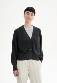 Schwarze Strickjacke mit V-Ausschnitt, geripptem Saum und fünf Knöpfen. Ausgestattet mit strukturierter Stoffqualität und langen Ärmeln, über einem hellgrauen Top getragen.