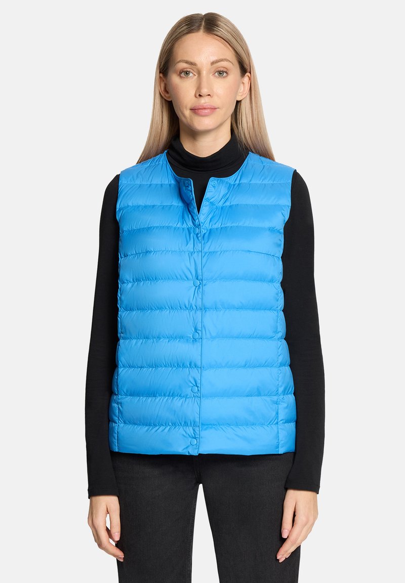 Frau trägt eine blaue, gesteppte, ärmellose Weste über einem schwarzen Langarmshirt und schwarzen Hosen, steht vor einem schlichten Hintergrund.