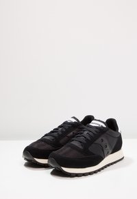 Black athletic sneakers with mesh, suede, and synthetic materials. Features include rounded toe, lace-up closure, and contrasting white midsole.
