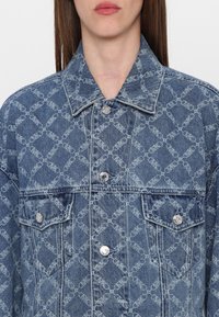 Kurtka jeansowa w jasnoniebieskim kolorze z powtarzającym się wzorem logo. Posiada kołnierz, kieszenie na piersi oraz srebrne guziki.