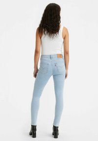 Mujer con cabello largo y rizado, vestida con una camiseta blanca sin mangas, jeans ajustados azul claro y botines negros con tacón, de pie de espaldas.