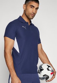 Námořnická modrá polo košile s bílými bočními akcenty, krátkými rukávy a stříbrným logem Puma. Model drží fotbal s grafickými detaily.