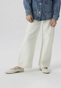 Pantalons blancs amples avec des motifs floraux brodés, associés à des chaussures Mary Jane de couleur claire. Portés avec une veste en denim.
