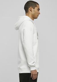 Weißer Hoodie mit Kängurutasche, Kordelzugkapuze und gerippten Bündchen. Glattes Material mit lässiger Passform, von der Seite betrachtet.