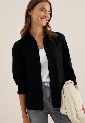 Zwarte button-up shirt met lange mouwen en een kraag, gecombineerd met een witte top en grijze jeans. Aan de ene kant wordt een lichte tas vastgehouden.