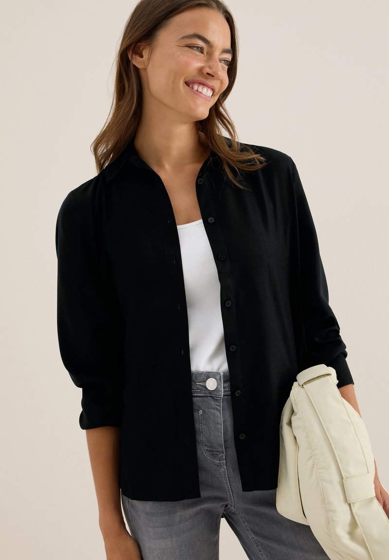 Chemise noire à manches longues avec col, associée à un top blanc et un jean gris. Un sac de couleur claire est tenu sur un côté.