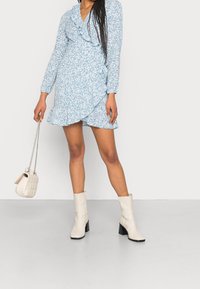 Kvinna iklädd en ljusblå blommig omslagsklänning med volanger, vita ankelskor och hållande en liten vit vadderad handväska med kedja.