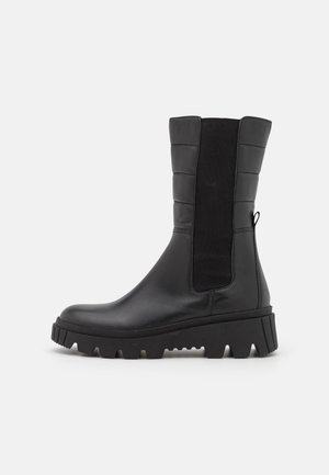 Botte noire en cuir mi-mollet avec semelle crantée épaisse, panneau latéral élastique, partie supérieure matelassée, et languette de traction sur fond blanc.