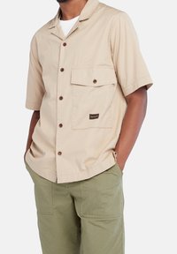 Beige korte mouwen overhemd met knoopsluiting en een camp collar, twee voorzakken en bruine knopen. Inclusief een Timberland-logo patch.