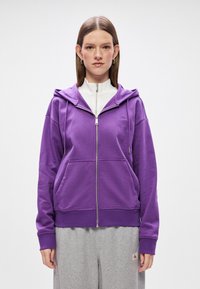 Fialová mikina na zips s kapucňou, prednými vreckami a rebrovanými manžetami. Nosená nad bielym topom s vysokým golierom, v kombinácii so šedými teplákmi.