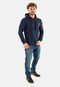 Felpa con cappuccio di colore blu navy, con zip frontale, tessuto strutturato e tasche laterali. Abbinata a jeans azzurri e scarpe scure con dettagli arancioni.