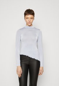 Lys blå langermet topp med mock neck og asymmetrisk kant; glatt stoff med bølgekant, kombinert med svarte tettsittende bukser.