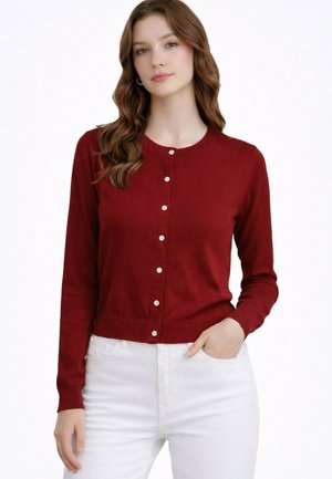 Femme aux cheveux bruns ondulés portant un cardigan rouge boutonné et un pantalon blanc, debout avec une main dans la poche sur fond blanc.