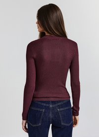 Ribbeliger bordeauxroter Pullover mit langen Ärmeln, rundem Halsausschnitt und taillierter Passform, kombiniert mit dunkelblauen Jeans mit Gesäßtaschen und Nähten.