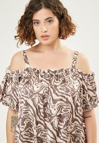 Bruine en crèmekleurige off-shoulder top met een gerimpelde hals en pofmouwen, voorzien van een abstract patroon. De stof lijkt lichtgewicht en soepel te zijn.