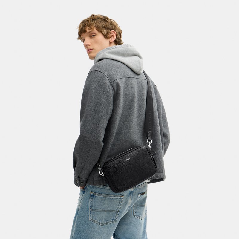 Joven con chaqueta gris y vaqueros azules lleva un bolso negro cruzado con una correa ancha sobre el hombro.